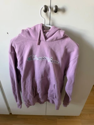 Ljuslila hoodie från Champion - Säljer en ljuslila hoodie från Champion med stor magficka och klassisk logga broderad på bröstet. Hoodien har huva med dragsko och är gjord i mjukt bomullsmaterial. Perfekt för en chill och avslappnad stil.