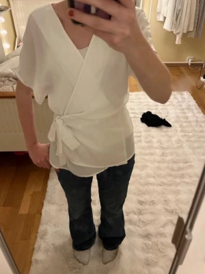 Vit omlottblus med knytband - Snygg vit omlottblus med korta ärmar och knytband i midjan. Blusen har en avslappnad passform och är tillverkad i ett lätt, luftigt tyg som ger en fräsch look. Perfekt att styla med jeans eller kjol för en trendig outfit.