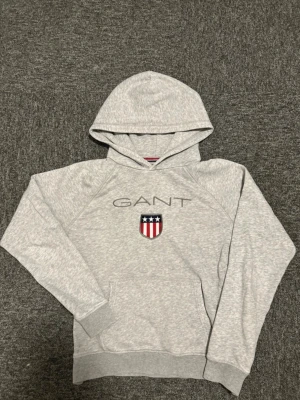 Grå hoodie från GANT  - Snygg ljusgrå hoodie från GANT med broderad logga och sköld på bröstet. Klassisk huva, ribbade muddar och magficka. Perfekt för en chill och sportig stil. Materialet är mjukt och bekvämt, troligen bomullsblandning.