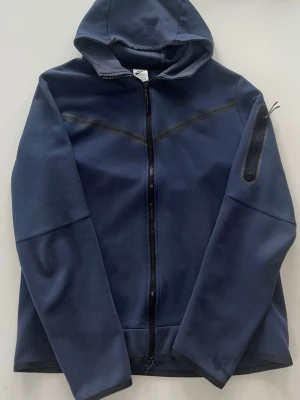 Mörkblå nike tech fleece - Snygg mörkblå hoodie från Nike med hel dragkedja framtill och huva. Jackan har en diskret ficka med dragkedja på ena ärmen och stilrena sömdetaljer över bröstet. Perfekt för en sportig och avslappnad look.