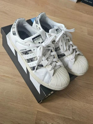 Adidas Superstar vita sneakers - Adidas Superstar sneakers – strl 37 1/3 Klassiska Adidas Superstar sneakers i vitt läder med svarta detaljer och mönstrade ränder. Skorna har den ikoniska snäckformade tån och vita skosnören – perfekta för en streetwear-look och passar till det mesta. Skick: Väl använda och slitna (se bilder för detaljer), därav priset. Storlek: 37 1/3 (Passar en storlek 36). 