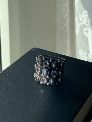 Bred silverring med färglada stenar - Unik bred ring i mörkt silver med flera infattade pärlor och lila stenar i olika storlekar. Ringen har en cool, bohemisk känsla och sticker verkligen ut på handen. Perfekt för dig som gillar statement-smycken och vill ha något annorlunda.