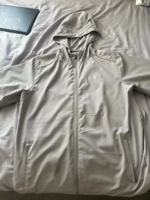 Ljusgrå vindjacka från Under Armour - Snygg ljusgrå vindjacka från Under Armour med huva och dragkedja framtill. Jackan har två sidofickor med dragkedja och är tillverkad i ett lätt, syntetiskt material som andas. Perfekt för dig som vill ha en sportig look. Jätte bra för detta väder just nu är inte så tjock och jätte fin tyg. Är 2 XL men hade sagt att den passar som XL också 