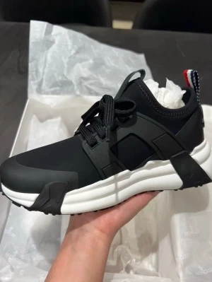 Svarta Moncler sneakers med vit sula - Snygga svarta sneakers från Moncler med chunky vit sula och svarta detaljer. Skorna har en sportig design med svarta snören och Moncler-logga bak. Röd, vit och blå flik baktill ger en extra touch. Tillverkade i en mix av textil och syntetmaterial. Om för kvitto!