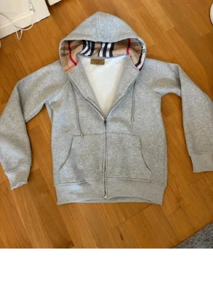 Grå zip-hoodie från Burberry - Snygg grå hoodie från Burberry med dragkedja och klassiskt rutigt foder i huvan. Mjuk bomullskänsla, två stora fickor framtill och justerbar huva med snören. Perfekt för en chill och stilren look.