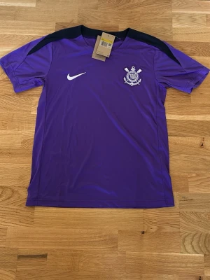 Lila Nike Corinthians träningströja - Snygg lila träningströja från Nike med Corinthians klubbmärke på bröstet och svart detalj över axlarna. Perfekt för fotbollsträning eller gymmet. Tröjan har korta ärmar och är tillverkad i ett lätt och ventilerande material.