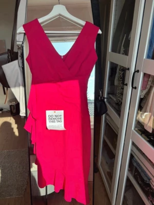 Rosa omlottklänning med volang - Snygg röd omlottklänning med v-ringning och volangdetaljer längs sidan och nederkanten. Klänningen är ärmlös och har en figurnära passform som framhäver siluetten. Perfekt för dig som vill sticka ut med en färgstark look. Materialet är stretchig. Helt ny och oanvänt. 
