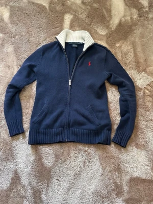 Ralphlouren zip hoodie  - Snygg marinblå ziptröja från Ralph Lauren med ribbstickade muddar och krage i kontrasterande vitt. Tröjan har två fickor framtill och en röd broderad logga på bröstet. Perfekt för en stilren och avslappnad look. Passar perfekt till 170-175 cm