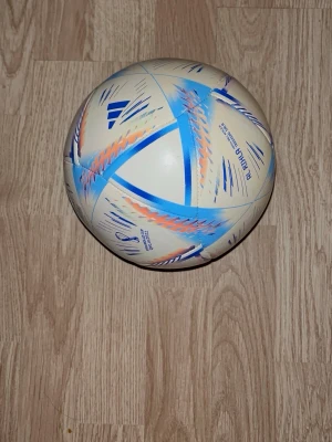 Adidas VM Qatar 2022 fotboll - Säljer en officiell Adidas fotboll från FIFA World Cup Qatar 2022. Bollen är vit med blå, orange och lila detaljer och har en modern design med tryckt turneringslogga. Perfekt för dig som älskar fotboll och vill ha en snygg boll att spela med eller samla på.