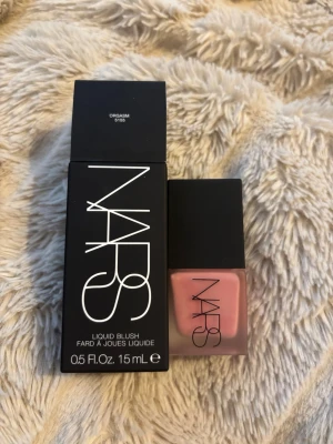 NARS Liquid Blush Orgasm 15ml - Flytande rouge från NARS i nyansen Orgasm, en ikonisk persikorosa färg med gyllene skimmer. Kommer i en stilren, frostad glasflaska med svart lock och tillhörande originalförpackning. Perfekt för att ge kinderna en fräsch och naturlig lyster.