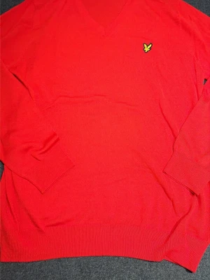 Röd stickad tröja från Lyle & Scott - Snygg röd stickad tröja från Lyle & Scott med V-ringning och klassisk gul logga på bröstet. Tröjan har ribbade muddar vid ärmslut och nederkant. Perfekt för dig som vill ha en enkel men stilren look.
