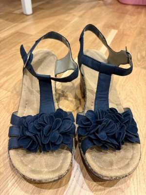Kilklackade sandaler med blomdetalj - Snygga blå (nästan svarta) sandaler med kilklack och dekorativa blommor framtill. Remmarna är i blått syntetmaterial och sulan har ett mönstrat korkutseende. Justerbart spänne vid ankeln för bra passform. Perfekta för en trendig sommarlook. Använda och väl omhändertagna. 