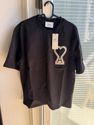 Svart t-shirt från Ami Paris - Cool svart t-shirt från Ami Paris med stort vitt broderat hjärta och A-logga på bröstet. Klassisk rund hals och korta ärmar. Materialet är mjukt och bekvämt, perfekt för en avslappnad look. Levereras i originalförpackning med Ami Paris-tryck.