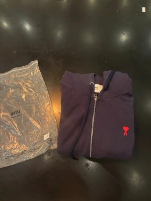 Mörkblå hoodie med dragkedja från Ami - Mörkblå hoodie från Ami med dragkedja framtill, huva med snören och två fickor. På bröstet finns Ami:s ikoniska röda hjärta med A. Tillverkad i mjukt bomullsmaterial, perfekt för en avslappnad och stilren look.