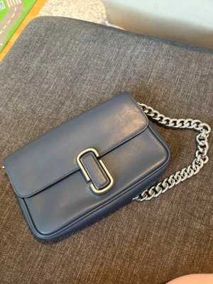Mörkblå handväska Marc Jacobs - Snygg mörkblå handväska från Marc Jacobs i slätt läder med silverfärgad kedja som axelrem. Väskan har en stilren rektangulär form och ett dekorativt spänne framtill. Perfekt för dig som vill ha en trendig accessoar med exklusiv känsla.