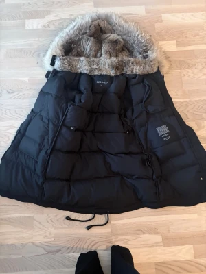 Svart dunjacka med fuskpäls från Cedrico - Säljer en svart dunjacka från Cedrico med stor huva och fuskpälsdetaljer. Jackan har dragkedja, knappar och flera fickor framtill. Perfekt för kalla vinterdagar och riktigt mysig med sin tjocka vaddering och fluffiga huva.