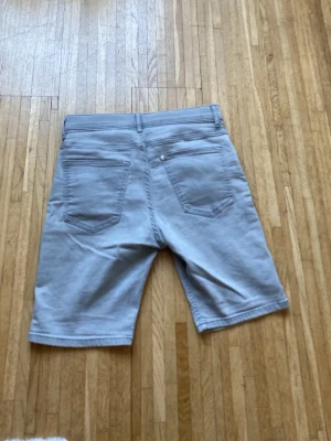 Ljusgråa jeansshorts med fem fickor - Snygga ljusgråa jeansshorts med klassisk femficksdesign och knappgylf. Modellen är rak och slutar strax ovanför knäna, perfekt för en avslappnad stil. Passar dig som gillar enkel och stilren look.