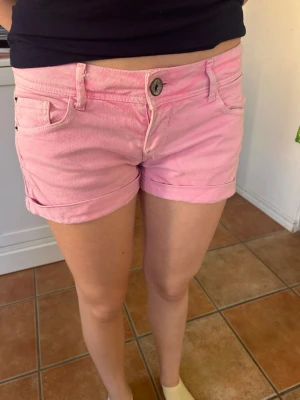 Rosa shorts från Clockhouse - Säljer ett par ljusrosa shorts från Clockhouse med klassisk femficksdesign och knappgylf. Shortsen har uppvikta benslut och bälteshällor, perfekta för varma dagar. Materialet är mjuk bomull som känns skönt mot huden.