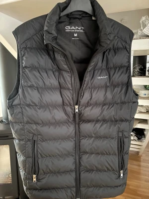 Svart dunväst från GANT, storlek M - Stilren svart dunväst från GANT med quiltad design och två dragkedjefickor. Västen har hög krage och dubbla dragkedjor framtill. Perfekt för lager-på-lager och snygg logga på bröstet. Materialet är lätt och håller värmen bra. Använd 3 gånger. 