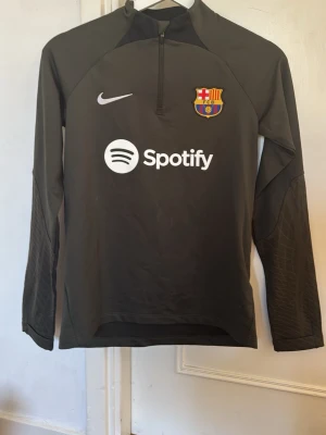 FC Barcelona träningströja Nike stl: L 147-158 cm - Snygg brun långärmad träningströja från Nike med FC Barcelona-logga och Spotify-tryck på bröstet. Tröjan har halv dragkedja framtill och är tillverkad i ett lätt och stretchigt material, perfekt för fotbollsträning eller gym.
