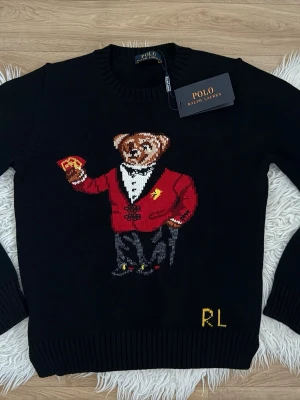 Svart stickad tröja Polo Ralph Lauren - Svart stickad tröja från Polo Ralph Lauren med ikonisk björn i röd kavaj på framsidan. Tröjan har rund halsringning, ribbstickade muddar och RL-broderi i gult nedtill. Perfekt för dig som gillar klassiska och lekfulla detaljer.
