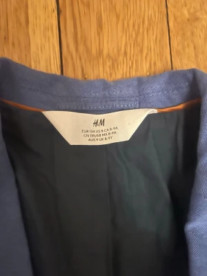 Blå kavaj från H&M barn - Snygg blå kavaj från H&M för barn, med två knappar framtill och klassiska fickor. Kavajen har en stilren passform och är helfodrad. Perfekt för dig som vill ha en enkel men stilren look. Materialet är troligtvis bomullsblandning.