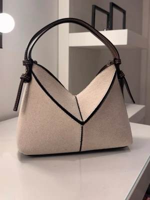 Zara väska - Säljer en stilren och trendig handväska från Zara i en fin kombination av beige tyg och bruna detaljer i konstläder. Väskan har en modern, strukturerad form som passar perfekt både till vardag och mer uppklädda tillfällen.  👜 Detaljer: * Kort handtag för en chic look * Justerbart axelband medföljer (kan bäras crossbody/över axeln) * Liten matchande necessär/plånbok ingår * Lätt men rymlig, får plats med det viktigaste * Neutrala färger som passar till allt  💫 Helt ny och oanvänd 📦 Skickar snabbt