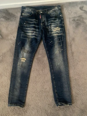 DSQUARED2 blå distressed jeans - Helt nya och snygga blå jeans från DSQUARED2. Byxorna har slitna detaljer, färgstänk och tryckt lönnlövsmotiv. 