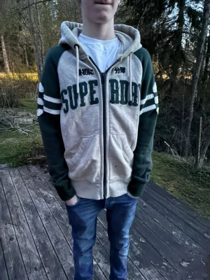 Superdry hoodie med dragkedja - Grå och mörkgrön hoodie från Superdry med dragkedja framtill och stora bokstäver på bröstet. Modellen på bilden är 182, skriv vid funderingar