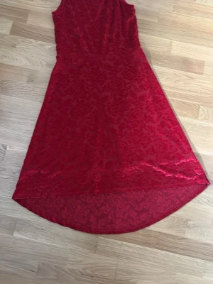 Röd ärmlös klänning med mönster - Snygg röd klänning med ärmlös design och subtilt blommigt mönster i tyget. Klänningen har en lätt utsvängd form och är något längre baktill. Perfekt för dig som vill sticka ut med en elegant look. Har 170 i storlek men är stor i storlek en så fungerar även som xs eller som liten s.