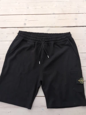 Svarta shorts från Stone Island XL - Helt nya shorts i storlek XL men passar garanterat till L (liten i storlek) Svarta shorts från Stone Island med elastisk midja och snörning. Klassisk logotyp på benet och praktiska fickor. Perfekta för en avslappnad stil och tillverkade i mjukt bomullsmaterial.