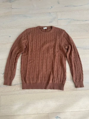 Brun kabelstickad tröja från John Henric  - Säljer en brun kabelstickad tröja med rund halsringning och ribbade muddar vid ärmslut och nederkant. Tröjan har ett klassiskt flätmönster och är perfekt för dig som gillar en stilren och mysig look.