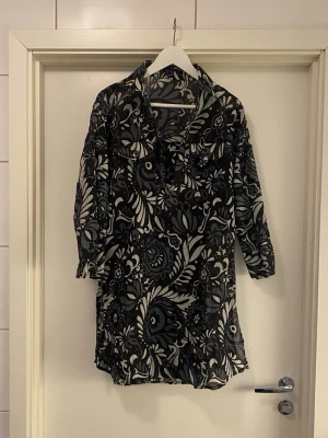 Skjortklänning / lång tunika - Fin skjortklänning / tunika med svart, blå och vitt blommönster, v-ringad med krage. Klänningen har trekvartsärm och två låtsas bröstfickor med knappdetaljer. Ärmar kan vikas upp och knäppas. Svalt material. Perfekt för dig som gillar en avslappnad men ändå snygg stil. Jättefint till vita byxor/ leggings👌
