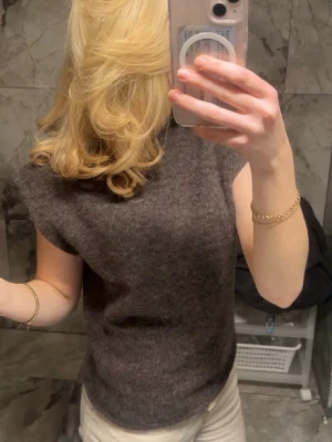 Mörkbrun ärmlös stickad tröja - Mysig mörkbrun stickad tröja med ärmlös design och rund hals. Perfekt för lager-på-lager och enkel att styla till olika outfits. Tröjan har en rak passform och är gjord i ett mjukt material som känns skönt mot huden.