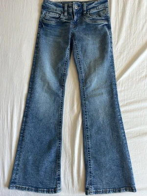 Blå bootcut jeans med slitningar - Snygga blå lågmidjade bootcut jeans med lätt slitna detaljer och klassiska fickor fram och bak. Jeansen har en utställd passform från knäna och är gjorda i denim med en cool vintage-look. Perfekta för dig som gillar retrostil och vill ha ett par jeans som sticker ut.