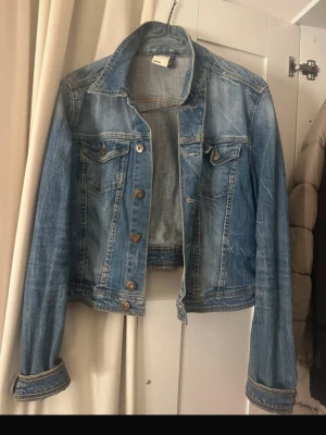 Klassisk blå jeansjacka - Snygg blå jeansjacka med slitningar och bruna knappar. Jackan har två bröstfickor med lock och knapp, samt klassisk krage. Perfekt till lager-på-lager och enkel att styla till olika outfits.