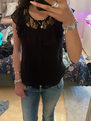 Svart blus med spetsdetaljer - Säljer en svart blus med transparent spets upptill och volangdetaljer vid axlarna. Blusen har knappar framtill och är ärmlös, vilket ger en luftig och elegant look. Perfekt att styla med jeans eller kjol för en trendig outfit.