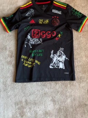 Ajax x Bob Marley Adidas tröja - Säljer en svart Ajax x Bob Marley limited edition tröja från Adidas med färgglada detaljer i rött, gult och grönt. Tröjan har tryck med Bob Marley, musiknoter, citat och Ziggo-logga. Materialet är lätt och andas, perfekt för fotboll eller streetwear.