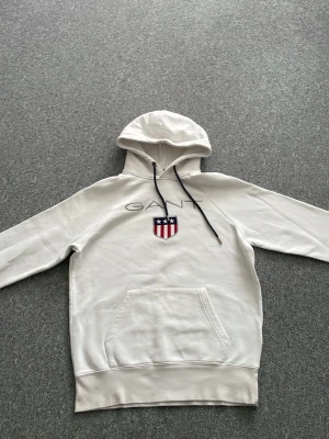 Vit hoodie från GANT med logga - Snygg vit hoodie från GANT med broderad logga och sköld på bröstet. Hoodien har huva med svarta dragsnören och en stor magficka framtill. Mjuk bomullskänsla och ribbade muddar vid ärmslut och nederkant. Perfekt för en avslappnad och stilren look.