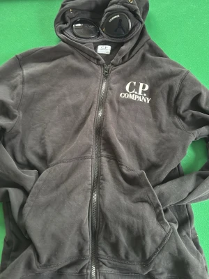 Svart hoodie med dragkedja från C.P. Company - Svart hoodie med dragkedja och huva från C.P. Company. Jackan har vita logotypdetaljer på bröstet och karakteristiska glasögon på huvan. Perfekt för en avslappnad streetwear-look. Säljer den pågrund av den är för liten skulle säga att den passar 170 storlek. Ny pris 2200 och jag säljer för 700