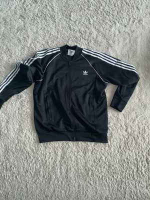  Adidas sst - Klassisk svart Adidas track jacket med dragkedja och vita ränder längs ärmarna. Tröjan har två fickor framtill och Adidas logga på bröstet. Perfekt för en sportig och avslappnad stil.