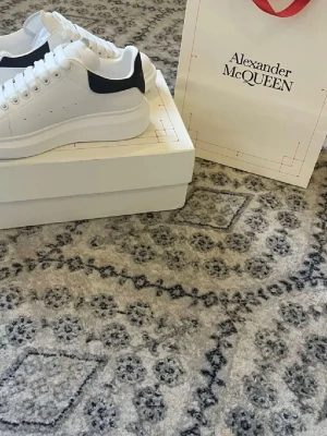 Vita Alexander McQueen sneakers - Stilrena vita sneakers från Alexander McQueen med chunky sula och svart detalj på hälen. Skorna har klassisk låg profil, vita skosnören och guldfärgad logga på plösen. Tillverkade i slätt läder för en clean och modern look. Obs låda medföljer ej
