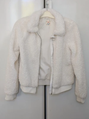 Vit teddyjacka med krage - Supermjuk och fluffig vit teddyjacka från HM med klassisk krage och dragkedja framtill. Jackan har ribbade muddar vid ärmslut och nederkant för extra komfort. Perfekt för dig som vill ha en mysig och trendig look. Längst ner på ena ribbade mudden lite mörka fläckar, bifogar bild. Jag kommer att stryka över min dotters namn som är skrivet på namnlappen under märkes och storleks lappen. Jag kommer att stryka över hennes namn med en mörk textilpenna