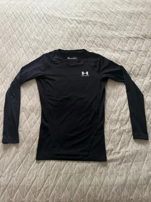 Svart Compression Shirt Under Armour - Svart långärmad träningströja från Under Armour med rund hals och vit logga på bröstet. Tillverkad i ett stretchigt och fukttransporterande material, perfekt för intensiva träningspass. Snygg och slimmad passform som sitter skönt på kroppen.