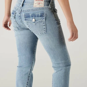 True Religion ljusblå bootcut jeans - Säljer ett par ljusblå jeans från True Religion med bootcut passform. Jeansen har klassiska kontrastsömmar, snygga fickdetaljer med lock och silverknappar bak samt en ikonisk patch i midjan. Perfekt för dig som gillar en avslappnad men trendig look.