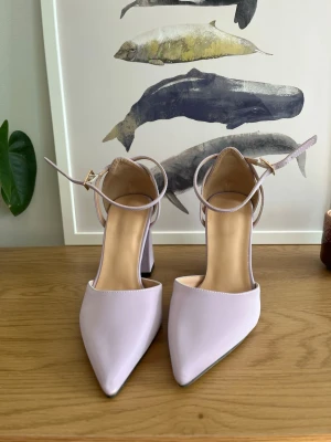 Ljuslila pumps med blockklack - Säljer ett par ljuslila pumps med bred blockklack och spetsig tå. Skorna har en elegant ankelrem med guldfärgat spänne och är tillverkade i ett slätt, matt material. Perfekta för dig som vill ha en trendig och stilren look. Se bilder på märken / fläckar. Köpt från SHEIN, säljs billigt 