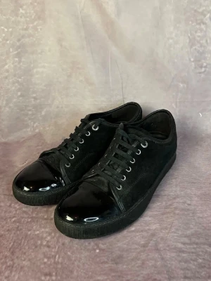 Svarta sneakers med lackad tå - Snygga helsvarta sneakers med ovandel i mocka och glansig lackad tå. Skorna har svarta skosnören, metallfärgade öljetter och en klassisk låg profil. Perfekta för dig som gillar stilrena och unika detaljer på dina sneakers.