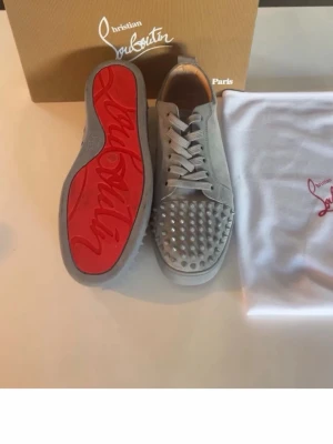 (Äkta) Christian Louboutin sneakers - Säljer ett par gråa sneakers från Christian Louboutin med klassisk röd sula och silverfärgade nitar på tån. Skorna är i mocka och har vita skosnören. Perfekta för dig som vill sticka ut med en lyxig och edgy stil.