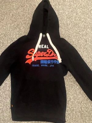 Svart Superdry hoodie med tryck - Svart hoodie från Superdry med stor logga i rött, vitt och blått framtill. Klassisk huva med vita snören och lång ärm. Perfekt för en chill och avslappnad stil.
