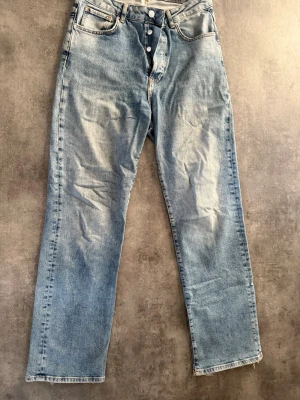 Ljusblå raka jeans med knappar - Säljer ett par ljusblå jeans med raka ben och knäppning framtill. Jeansen har klassiska fem fickor och råa, fransiga benslut för en avslappnad look. Tillverkade i denim med en skön vintagekänsla. Har ett litet hål vid inre låret, men syns inte vid användning